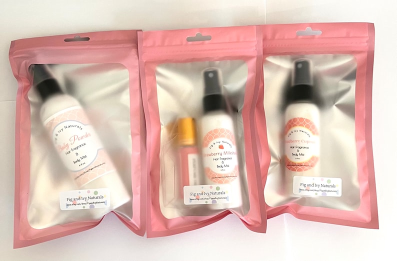 May include: Three pink-packaged body mists and hair fragrances. The bottles are white with black spray nozzles and labels that read "Baby Powder", "Strawberry Milkshake", and "Strawberry Cupcake". Each package has the "Fig and Ivy Naturals" logo.