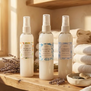 May include: Three clear linen spray bottles with white spray tops and labels. The labels read "Sunlit Citrus", "Coastal Calm", and "Cabana Breeze". The bottles are on a wooden surface with lavender and a bowl of stones.