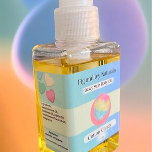 May include: A clear, square bottle of "Fig and Ivy Naturals" "Dewy Skin Body Oil" with a white pump dispenser. The label features a cotton candy graphic and ingredients list. The oil is a golden yellow color.