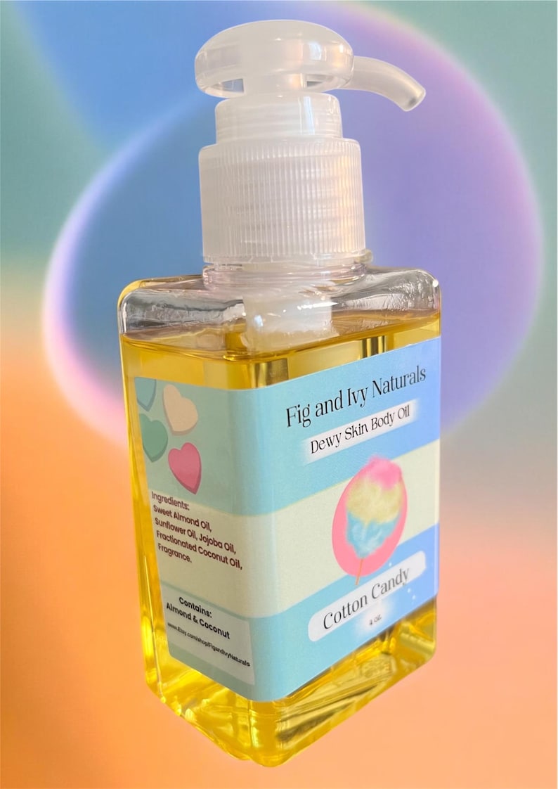 May include: A clear, square bottle of "Fig and Ivy Naturals" "Dewy Skin Body Oil" with a white pump dispenser. The label features a cotton candy graphic and ingredients list. The oil is a golden yellow color.