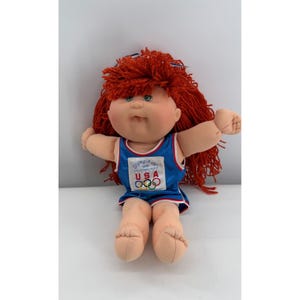 May include: A doll with bright red yarn hair wearing a blue athletic tank top with white trim. The shirt features the text Olympikids 1996 Official Team Mascot USA and the five-ring Olympic logo. The doll has a light skin tone and green eyes.