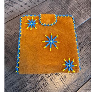 May include: A square tan suede pouch featuring a flap closure with a snap button. The edges and the flap are trimmed with alternating blue and yellow beads. Three star-shaped patterns made of blue and yellow beads decorate the front surface.