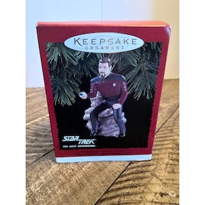 Könnte beinhalten: Eine Hallmark Keepsake Ornament-Schachtel mit einer Commander William Riker-Figur aus Star Trek The Next Generation. Die Verpackung zeigt die Figur in einer kastanienbraunen und schwarzen Uniform, hockend auf einem Felsen vor einem Tannenbaum.