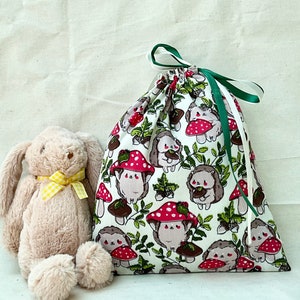 Pode incluir: Uma bolsa de tecido branco com cord&atilde;o de ajuste verde. A bolsa &eacute; decorada com um padr&atilde;o repetido de cogumelos vermelhos e ouri&ccedil;os.