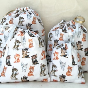 May include: Four white drawstring bags featuring a pattern of playful kittens in various poses. The bags are made from cotton fabric and have white ribbon ties.