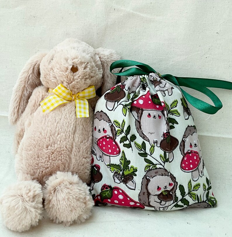 Pode incluir: Um brinquedo de pel&uacute;cia de coelho bege com um la&ccedil;o amarelo fica ao lado de uma bolsa de cord&atilde;o de tecido verde e branco com uma estampa de ouri&ccedil;o e cogumelos.