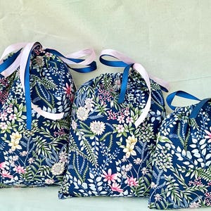 May include: Three blue drawstring bags with pink and blue ribbon ties. The bags are made of fabric with a floral pattern in shades of pink, white, and green.