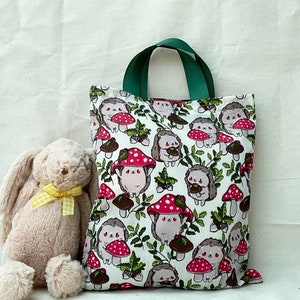 Pode incluir: Uma bolsa tote verde e branca com um design de cogumelos e ouri&ccedil;os. A bolsa tem uma al&ccedil;a de lona verde e um fundo branco com detalhes marrons, vermelhos e verdes.