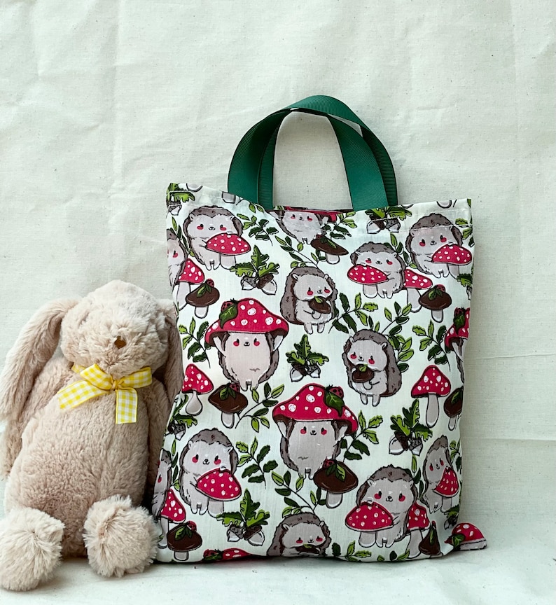 Pode incluir: Uma bolsa tote verde e branca com um design de cogumelos e ouri&ccedil;os. A bolsa tem uma al&ccedil;a de lona verde e um fundo branco com detalhes marrons, vermelhos e verdes.
