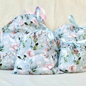 May include: Four blue drawstring bags with a floral print featuring pink roses, green leaves, and white hummingbirds. The bags are made of fabric and have a light blue background.