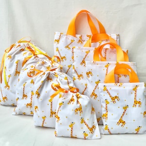 Pode incluir: Um conjunto de seis sacos de tecido branco com estampa de girafa amarela e laços de fita laranja. Os sacos são de vários tamanhos e estilos, incluindo totes e sacos de cordão.