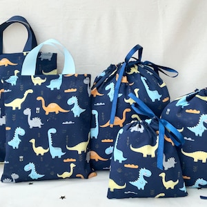 Pode incluir: Conjunto de cinco sacos de lona azul com um padrão de dinossauro. Os sacos são de diferentes tamanhos e estilos, incluindo sacos de mão e sacos de cordão.