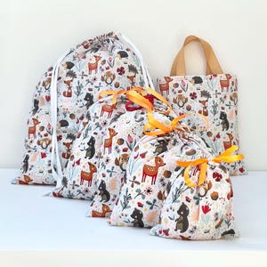 May include: A selection of drawstring bags and a tote bag, all patterned with woodland creatures. The bags feature a white backdrop with illustrations of deer, foxes, hedgehogs, and bears. The drawstring bags have white cords and orange ribbons.