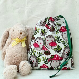 Pode incluir: Uma bolsa de tecido branco com cord&atilde;o de ajuste verde. A bolsa tem um padr&atilde;o de cogumelos vermelhos e ouri&ccedil;os castanhos sobre um fundo branco. Um coelho de pel&uacute;cia castanho est&aacute; sentado na frente da bolsa.