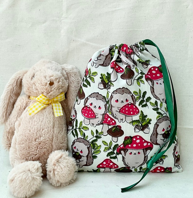 Pode incluir: Uma bolsa de tecido branco com cord&atilde;o de ajuste verde. A bolsa tem um padr&atilde;o de cogumelos vermelhos e ouri&ccedil;os castanhos sobre um fundo branco. Um coelho de pel&uacute;cia castanho est&aacute; sentado na frente da bolsa.