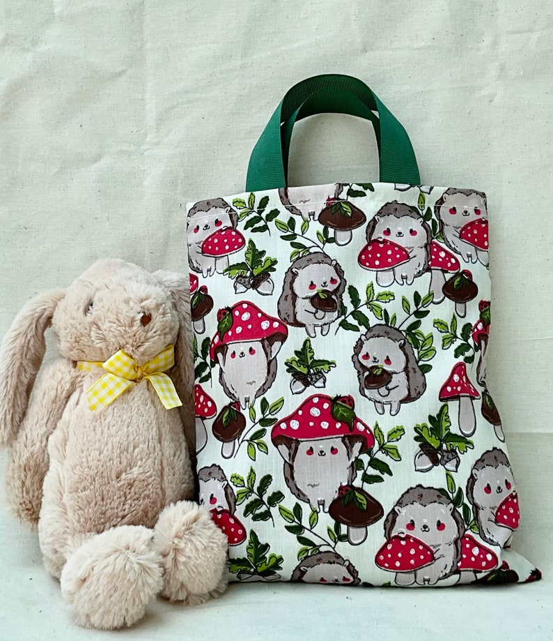 Pode incluir: Uma bolsa de tecido verde e branca com um padr&atilde;o repetido de ouri&ccedil;os e cogumelos. Os ouri&ccedil;os est&atilde;o segurando cogumelos e os cogumelos s&atilde;o vermelhos com bolinhas brancas.