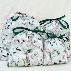 Pode incluir: Quatro sacos florais com cordão em vários tamanhos. Os sacos apresentam um fundo verde claro com flores rosa e brancas e folhas verdes. Cada saco é amarrado com uma fita verde ou rosa.