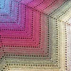 Op de afbeelding: Een gehaakt deken met een kleurverloop van blauw naar roze, groen en beige. Het deken heeft een kantpatroon met open vierkanten.