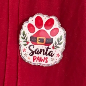 May include: A circular pin featuring a red paw print design with a black belt and gold buckle across the center. The text "Santa PAWS" is printed in black script below the belt, surrounded by green holly leaves and red berries on a white background with a silver glitter border.