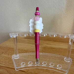 Op de afbeelding: Een magenta glitterende balpen met een decoratieve topper in een witte geschulpte vorm en een stapel kralen in roze, wit en geel, geplaatst in een doorzichtige acryl displaystandaard met meerdere sleuven voor het organiseren van pennen.