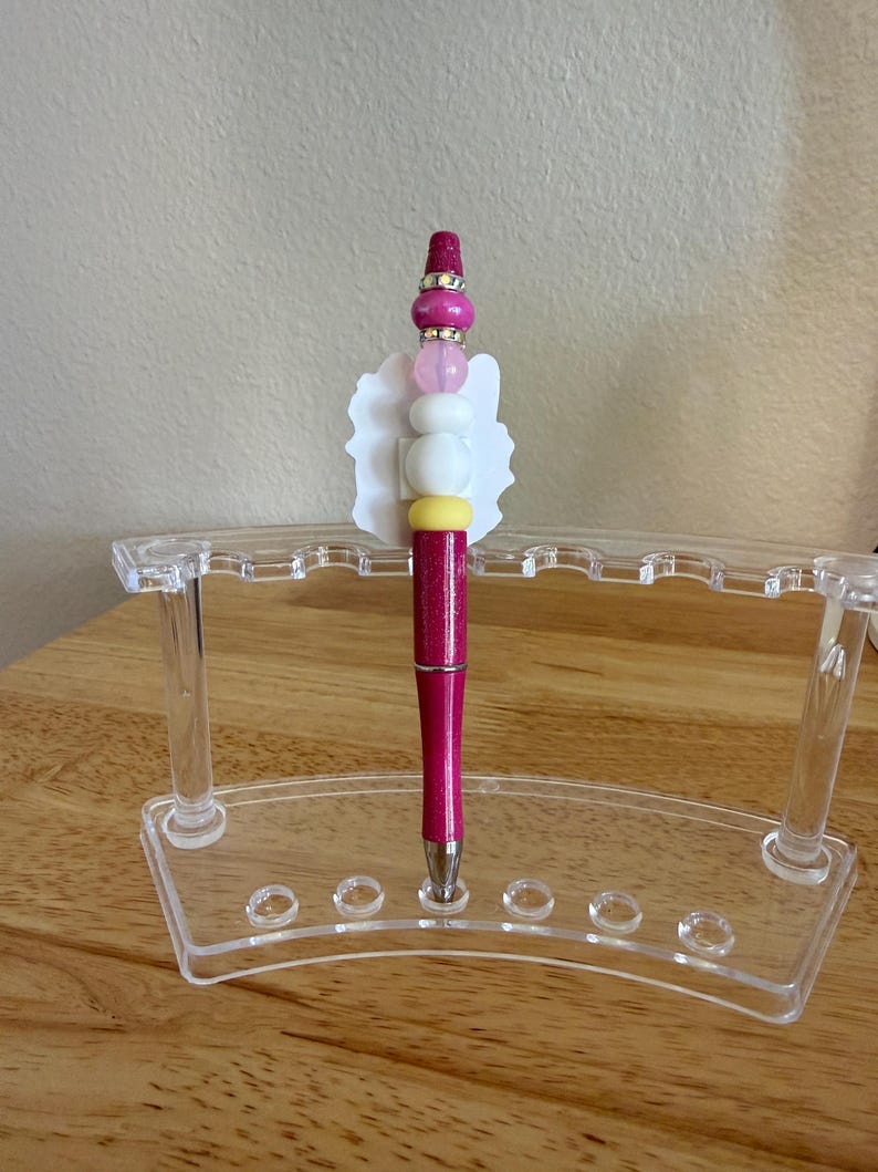 Op de afbeelding: Een magenta glitterende balpen met een decoratieve topper in een witte geschulpte vorm en een stapel kralen in roze, wit en geel, geplaatst in een doorzichtige acryl displaystandaard met meerdere sleuven voor het organiseren van pennen.