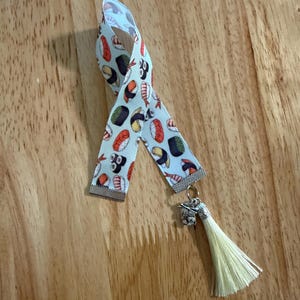 May include: Sushi-themed bookmark with a light blue ribbon patterned with various sushi rolls. The bookmark has a silver-colored charm and a cream-colored tassel, perfect for any book lover.
