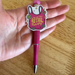 Op de afbeelding: Een magenta glitterbalpen met een decoratieve topper in de vorm van een roze boodschappentas met de tekst "TIME FOR THAT RETAIL THERAPY", omringd door kleine bloemillustraties en geaccentueerd met een glanzende roze kraal en zilveren ringen.
