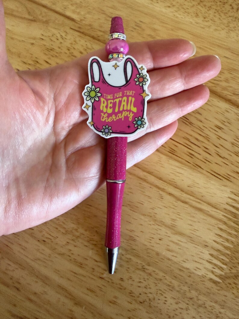 Op de afbeelding: Een magenta glitterbalpen met een decoratieve topper in de vorm van een roze boodschappentas met de tekst "TIME FOR THAT RETAIL THERAPY", omringd door kleine bloemillustraties en geaccentueerd met een glanzende roze kraal en zilveren ringen.