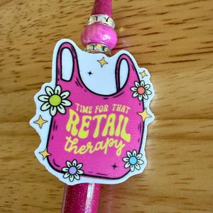 Op de afbeelding: Een roze glitterpen met een decoratieve hanger in de vorm van een boodschappentas. De hanger bevat de tekst "TIME FOR THAT RETAIL therapy" in het geel, omringd door kleine bloem- en sterillustraties, met een roze kraal en strass-steentjes.