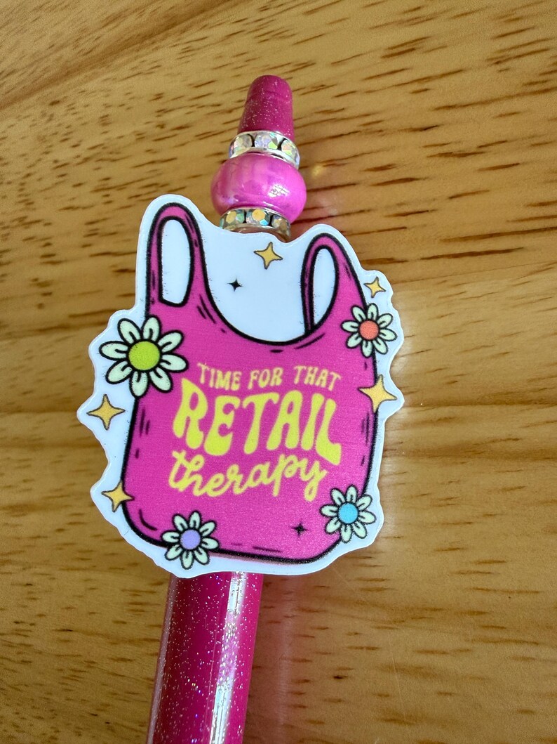 Op de afbeelding: Een roze glitterpen met een decoratieve hanger in de vorm van een boodschappentas. De hanger bevat de tekst "TIME FOR THAT RETAIL therapy" in het geel, omringd door kleine bloem- en sterillustraties, met een roze kraal en strass-steentjes.