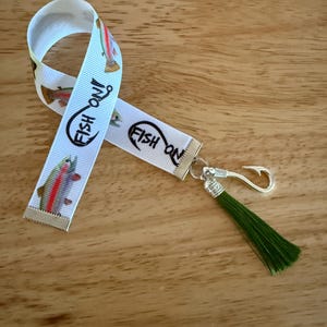 Puede incluir: Llavero de cinta blanca con diseño de pez y anzuelo. La cinta tiene las palabras "FISH ON" y un gráfico de pez. Un gancho plateado y una borla verde están unidos a la cinta.