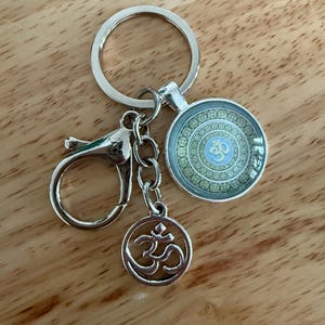 Puede incluir: Llavero plateado con un colgante redondo con un diseño de mandala azul y verde y el símbolo Om. El llavero incluye un mosquetón y un dije más pequeño con el símbolo Om.