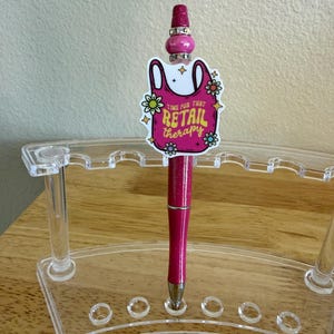 Op de afbeelding: Een glitterende roze pen met een decoratieve topper in de vorm van een boodschappentas met de tekst "TIME FOR THAT RETAIL THERAPY" omringd door kleine bloemen. De pen is versierd met zilverkleurige kralen en staat in een doorzichtige houder.