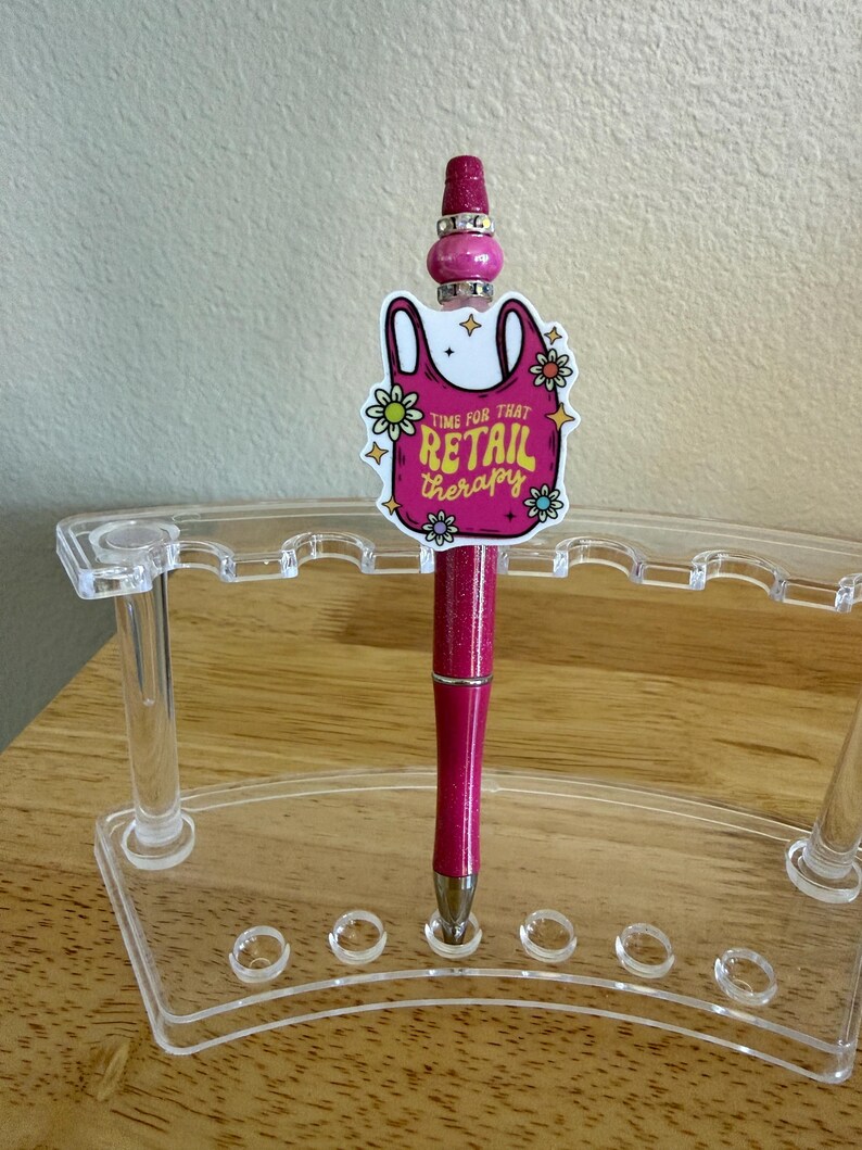 Op de afbeelding: Een glitterende roze pen met een decoratieve topper in de vorm van een boodschappentas met de tekst "TIME FOR THAT RETAIL THERAPY" omringd door kleine bloemen. De pen is versierd met zilverkleurige kralen en staat in een doorzichtige houder.