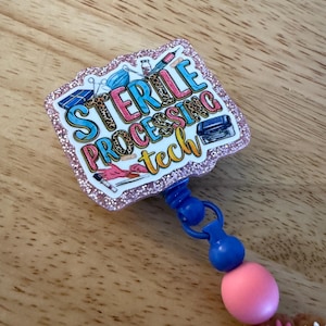 Op de afbeelding: Een badge reel met een roze glitterrand en de woorden "STERILE PROCESSING tech" in kleurrijke letters. De badge heeft medische illustraties en een blauw en roze kralenaccent.