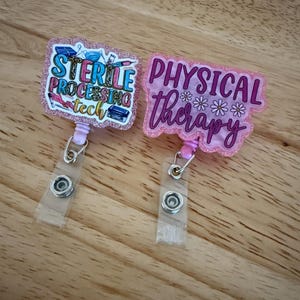 May include: Two badge reels with glitter detailing. One reads "STERILE PROCESSING tech" in teal, pink, and yellow. The other says "PHYSICAL therapy" in pink with white flower accents. Both have clear plastic straps and silver metal clips.