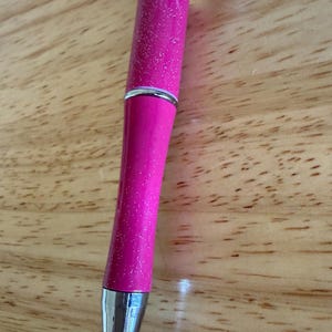 Op de afbeelding: Een intrekbare balpen met een felroze glitterbehuizing en een zilverkleurige metalen punt. De bovenkant van de pen is voorzien van een decoratieve roze topper met een grafisch ontwerp van blauwe en witte bloemen en een zwart sterretje.