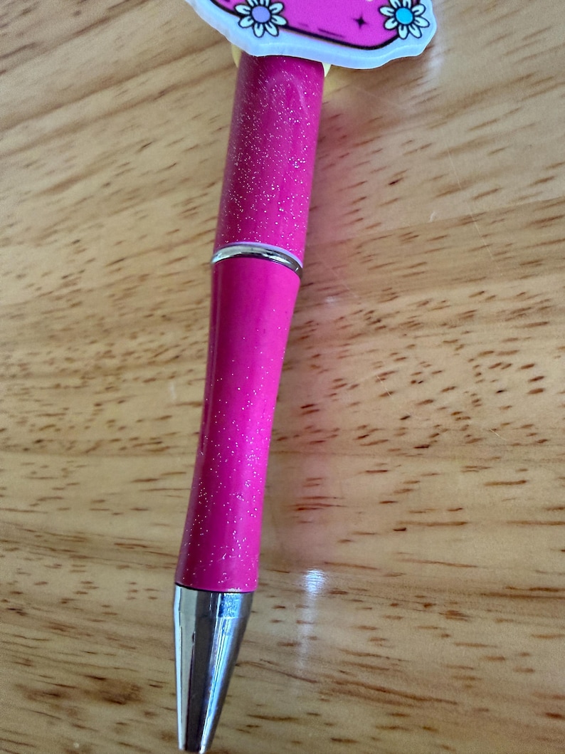 Op de afbeelding: Een intrekbare balpen met een felroze glitterbehuizing en een zilverkleurige metalen punt. De bovenkant van de pen is voorzien van een decoratieve roze topper met een grafisch ontwerp van blauwe en witte bloemen en een zwart sterretje.