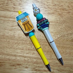 May include: Two pens with decorative toppers. One pen is yellow with a "Stool Sample" sticker. The other pen is white with a charm that says "Coffee Scrubs Rubber Gloves". Both pens have silver accents and black tips.