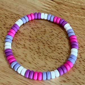 May include: A colorful bracelet made of small, flat, round beads. The beads are arranged in a circular pattern, featuring a mix of white, pink, purple, and lavender colors. The bracelet is displayed on a wooden surface.