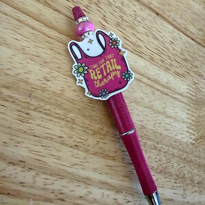 Op de afbeelding: Een roze glitterbalpen met een decoratieve topper in de vorm van een boodschappentas met de tekst "TIME FOR THAT RETAIL THERAPY" omringd door kleine bloemillustraties, geaccentueerd met een roze kraal en twee zilverkleurige strass-ringen.
