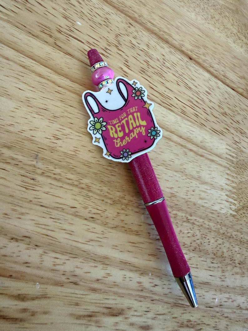 Op de afbeelding: Een roze glitterbalpen met een decoratieve topper in de vorm van een boodschappentas met de tekst "TIME FOR THAT RETAIL THERAPY" omringd door kleine bloemillustraties, geaccentueerd met een roze kraal en twee zilverkleurige strass-ringen.
