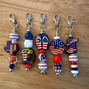Puede incluir: Una colección de llaveros con temas patrióticos. Cada llavero presenta un cierre plateado y una variedad de dijes, incluyendo una bandera de Texas, una bota de vaquero, chanclas y una rosquilla, todos adornados con diseños rojos, blancos y azules.
