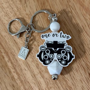 May include: A silver keychain with a charm shaped like an eye exam machine, with the words "one or two" above it. A small charm with an eye chart is also attached. The keychain is on a wooden surface.