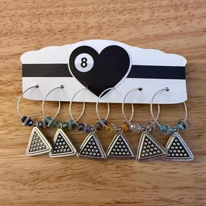 Puede incluir: Juego de seis colgantes de vino plateados. Cada colgante presenta un colgante triangular con cuentas de colores variados. Los colgantes cuelgan de un aro plateado. Detrás de los colgantes hay una tarjeta en blanco y negro con un corazón y el número 8.