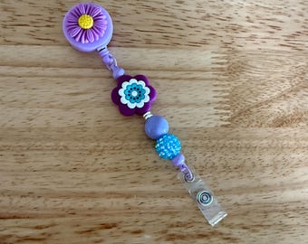 May include: A purple flower-shaped badge reel with a yellow centre, connected to a beaded strand with a turquoise flower and blue beads. The reel has a clear plastic clip for attaching to clothing.