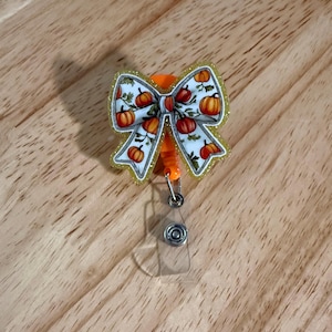 May include: A badge reel with a bow-shaped design featuring orange pumpkins on a white background. The bow has a gold glitter outline and is attached to an orange reel with a clear plastic clip.