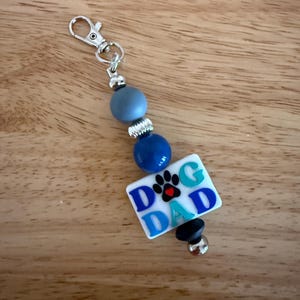 Op de afbeelding: Een sleutelhanger met een zilveren sluiting, blauwe kralen en een witte rechthoekige bedel. De bedel heeft de woorden "DOG DAD" in blauw, een pootafdruk en een kleine zwarte kraal. De achtergrond is een houten oppervlak.