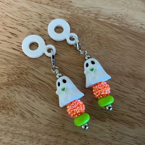 Op de afbeelding: Een paar Halloween-oorbellen. Elke oorbel heeft een witte spookbedel met zwarte ogen en een groene mond, gecombineerd met oranje en groene kralenaccenten. De oorbellen hebben zilverkleurige haken.