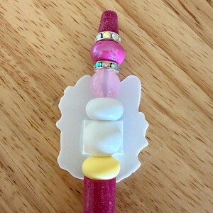Op de afbeelding: Een pen met een donkerroze glitterhouder, bekroond met een stapel kralen waaronder een donkerroze glitterkraal, twee zilverkleurige strass-tussenstukjes, een lichtroze kraal, twee witte kralen en een gele kraal, met een platte witte uitgesneden vorm.